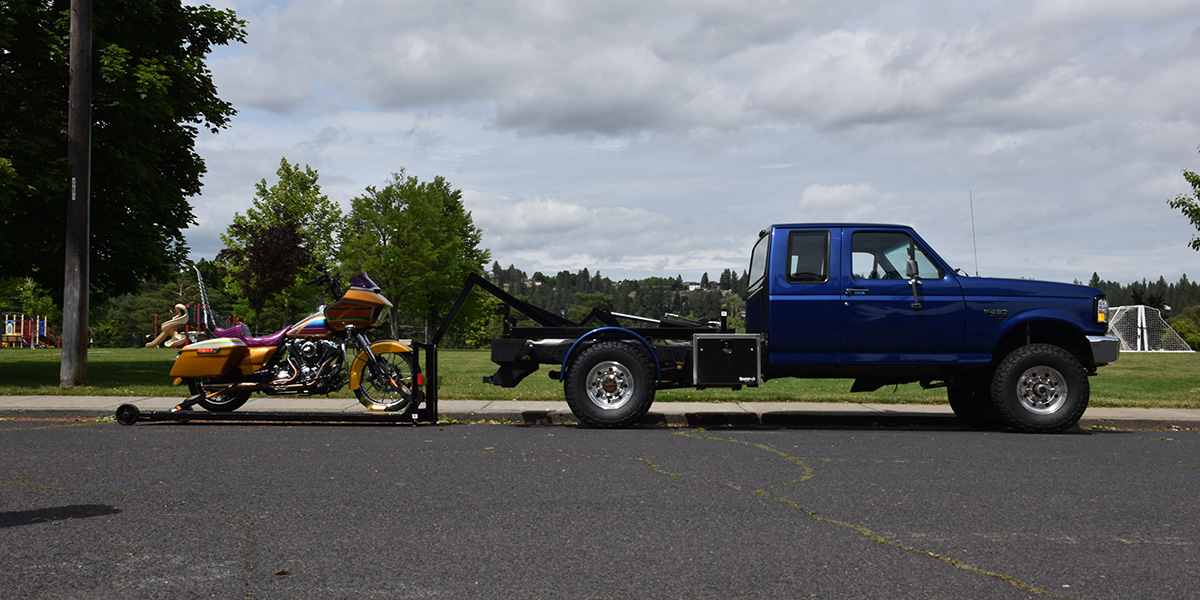 Royal blue flatbed tow truck loaded with a secured motorcycle on a Northwest highway, ready for transport.